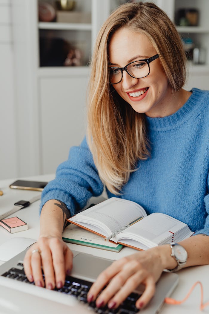 gallery-6 Smiling woman in eyeglasses typing on laptop at desk with open book.