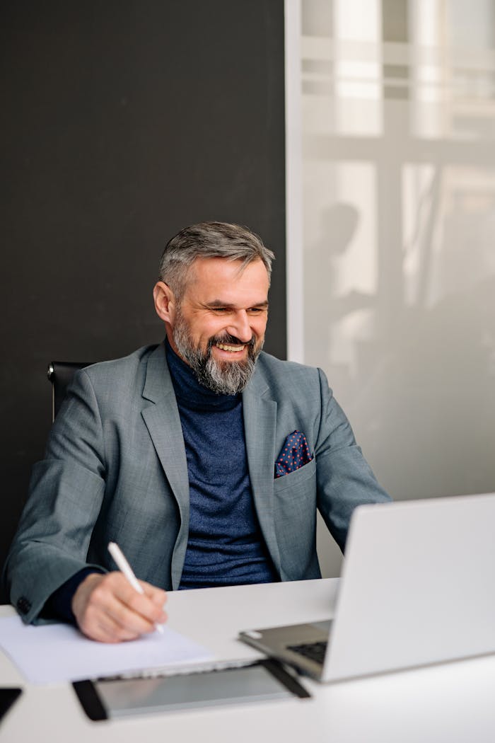Businessman in gray suit smiling confidently while working in a modern office environment. Indoors, natural light.