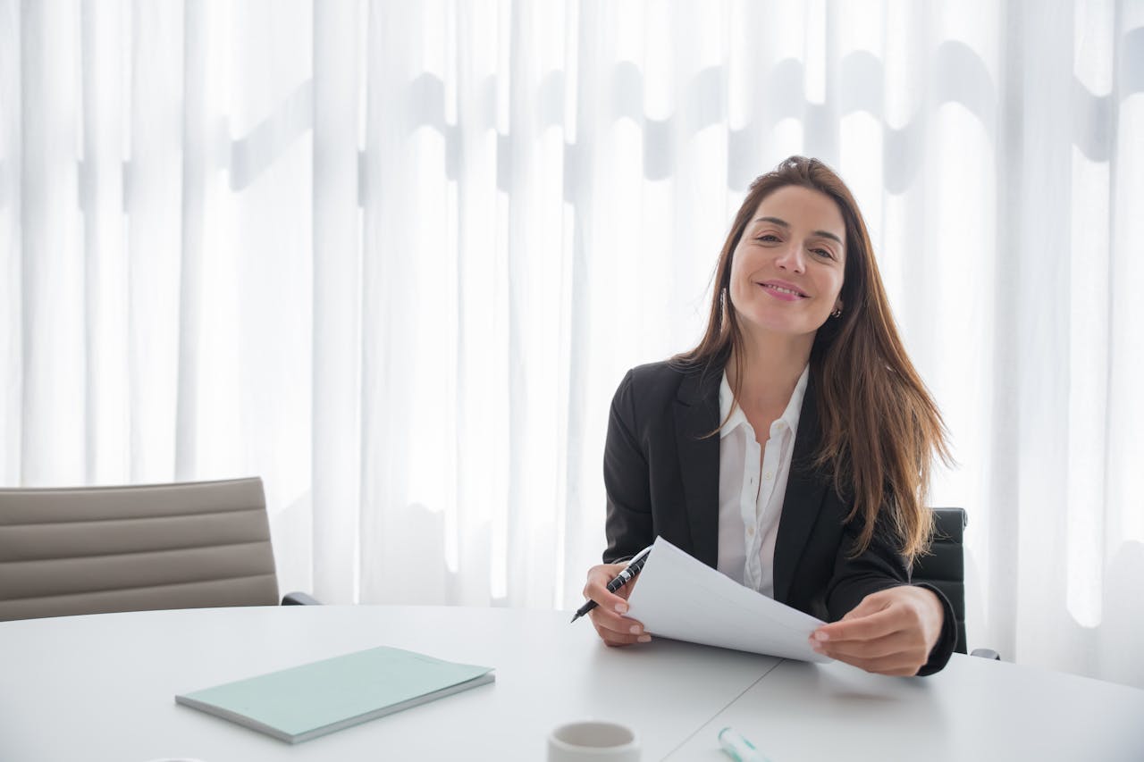 gallery-4 Smiling businesswoman in a blazer holding paper at an office table indoors.